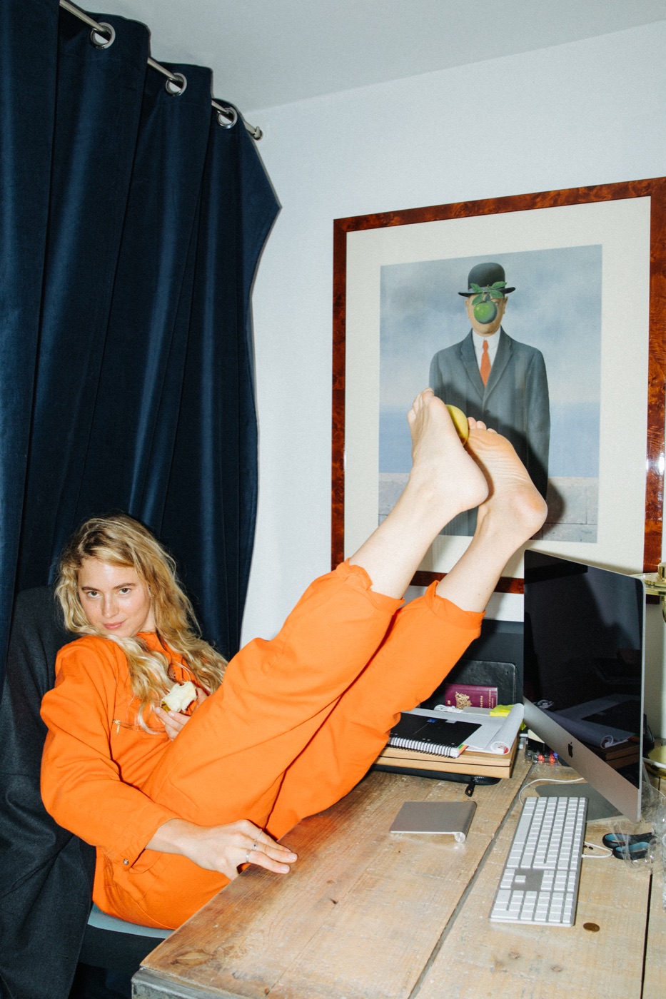 Marije at her desk in an orange jumpsuit, feet up in front of a Magritte print